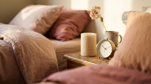 A serene bedroom corner with soft natural linen bedding, a small clock and a candle in gentle evening light
